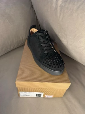 Svarta Louboutin sneakers - Säljer ett par svarta sneakers från Christian Louboutin med mattsvarta nitar på tån och klassisk röd sula. Skorna är i mocka och har svarta skosnören. Perfekta för dig som vill sticka ut med en edgy och lyxig stil.