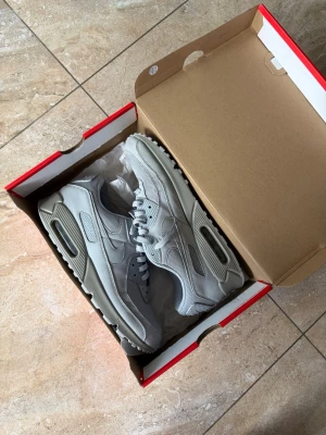 Nike Air Max 90 grå sneakers - Snygga Nike Air Max 90 sneakers i helgrå färg med klassisk siluett. Skorna har ovandel i mesh och läder, synlig Air Max-dämpning i sulan och diskreta Nike-loggor på sidorna. Perfekta för dig som gillar stilrena och sportiga sneakers.