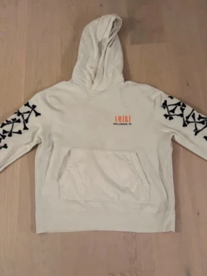 Vit hoodie från Amiri med tryck - Snygg vit hoodie från Amiri med svart benmönster längs båda ärmarna och orange Amiri-tryck på bröstet. Klassisk känguruficka framtill och huva. Perfekt för dig om som gillar streetwear och vill sticka ut lite extra.