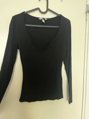 Svart ribbad v-ringad topp - Snygg svart ribbad topp med v-ringning och långa ärmar. Figurnära passform som framhäver siluetten. Perfekt att matcha med jeans eller kjol för en trendig look.