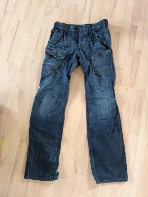 G-STAR jeans 31/38 - Säljer ett par blå jeans från G-Star RAW med flera stora fickor framtill och bak. Jeansen har synliga sömmar, knappar i gylfen och förstärkta partier vid knäna. Perfekt för dig som gillar en lite tuffare streetstil.