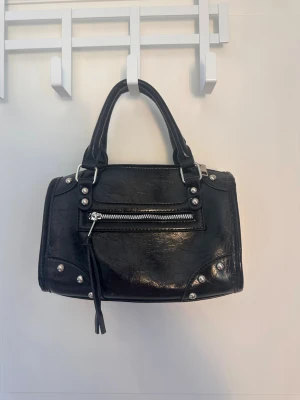 Svart handväska med nitar och dragkedja - Cool svart handväska i skinnimitation med silverfärgade nitar och detaljer. Väskan har dubbla handtag, en dragkedja framtill och dekorativa sömmar. Perfekt för dig som gillar en edgy och trendig look.