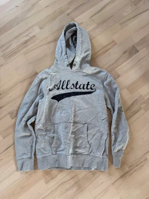 Grå hoodie Allstate med tryck - Snygg grå hoodie från Allstate med svart tryck på bröstet och texten 'Allstate'. Klassisk känguruficka framtill, ribbade muddar och huva med knappar vid halsen. Perfekt för en avslappnad streetstil.