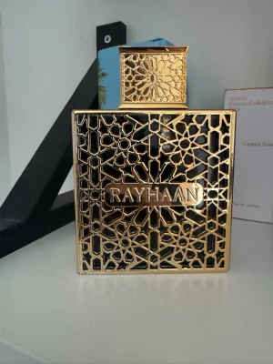 Rayhaan Elixir parfym i guldig flaska - Rayhaan Elixir parfym i en lyxig, fyrkantig flaska med guldig metallisk dekor och orientaliskt mönster. Flaskan har en svart bakgrund och ett detaljerat guldlock. Perfekt för dig som gillar exklusiva och unika dofter.