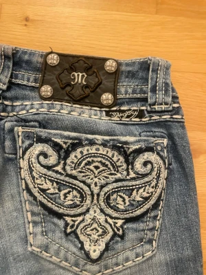Miss Me bootcut jeans stl 26 - Snygga blå bootcut jeans från Miss Me med slitningar på låren och dekorativa detaljer på bakfickorna. Jeansen har ljus tvätt, kontrastsömmar och läderpatch med metallnitar bak i midjan. Perfekta för en trendig och avslappnad look. Använda fåtal gånger. Pris kan diskuteras Kostar ca  2300 nya.