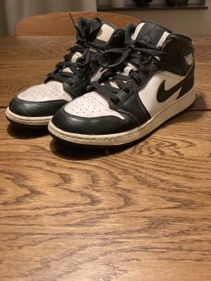 Nike Air Jordan 1 svartvita sneakers - Säljer ett par Nike Air Jordan 1 sneakers i svart och vitt läder med klassisk Swoosh-logga på sidan och vingar på ankeln. Skorna har högt skaft och perforerad tå för extra ventilation. Säljer för det är för små för mig nu 