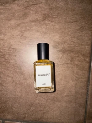 Lush Vanillary parfym - En stilren parfymflaska från Lush med doften Vanillary. Flaskan är rektangulär i genomskinligt glas med svart lock och vit etikett. Innehållet är ljusgult och flaskan har en modern, minimalistisk design.