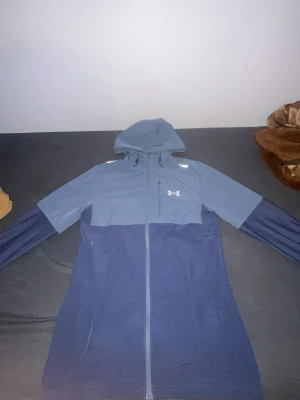 Blå vindjacka från Under Armour - Snygg tvåfärgad blå vindjacka från Under Armour med huva och hel dragkedja. Jackan har reflekterande detaljer på axlarna och logga på bröstet. Perfekt för blåsiga dagar och sportiga outfits. Tillverkad i lätt och slitstarkt material.