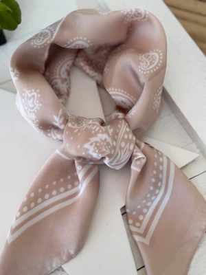 Champagnefärgad scarf i silkeslook – couture paisley  - Elegant scarf i skimrande champagne-ton med sofistikerat paisleymönster och exklusiv satinfinish. Den mjuka silkeslooken tillsammans med de ton-i-ton inspirerade detaljerna ger en tidlös couture-känsla med understated luxury-vibe.  Den varma champagnefärgen skiftar vackert i ljuset och gör scarfen lika chic till en minimalistisk outfit som stylad runt halsen, i håret eller på väskan.  70 x 70 cm. En feminin och exklusiv statement-accessoar med modern vintagekänsla. ✨