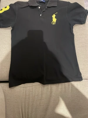 Svart pikétröja från Ralph Lauren - Svart pikétröja från Ralph Lauren med stor gul logga på bröstet och gul siffra på ärmen. Klassisk krage och knäppning framtill. Perfekt för en stilren och sportig look.