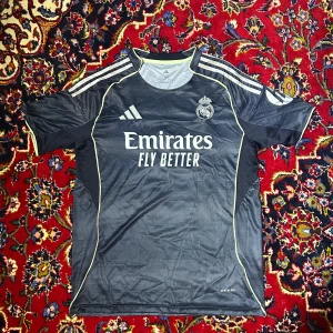 Real Madrid 25/26 Away Tröja - Snygg Real Madrid fotbollströja från Adidas i storlek L, med Mbappé och nummer 9 på ryggen. Tröjan är mörkblå med tunna ljusa ränder, vita detaljer och gula inslag vid krage och sömmar. Klassisk rund hals och korta ärmar, tillverkad i lätt funktionsmaterial.