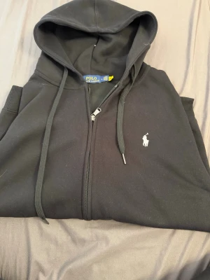 Svart zip hoodie från Polo Ralph Lauren - Klassisk svart hoodie med dragkedja från Polo Ralph Lauren. Hoodien har huva med dragsko, ribbade muddar och fickor framtill. Ikoniska vita loggan broderad på bröstet. Tillverkad i mjuk bomullsblandning för skön känsla.