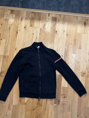 Mörkblå cardigan från Moncler - Snygg svart ziptröja från Moncler med ståkrage och två fickor framtill. Tröjan har en vit och röd rand på ena ärmen samt Moncler-logga på bröstet. 