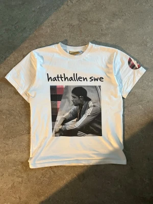 Vit t-shirt från Hatthallen SWE - Vit t-shirt från Hatthallen SWE med tryck framtill av en person i sportkläder och texten 'hatthallen swe'. Rund halsringning och korta ärmar. Tryckdetalj även på ena ärmen. Perfekt för en streetwear-look. 5 cold wash tvättar och inga slitningar. Kom gärna med bud