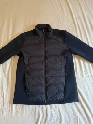 Sail Racing Cardigan - Snygg svart cardiganjacka från Sail Racing med quiltad framsida och släta stickade ärmar. Jackan har hög krage, dragkedja framtill och diskret Sail Racing-logga på ärmen. Perfekt för lager-på-lager och stilrena outfits. Detta är den nya modellen köpt från Collection of Brands. Nypris: 3000 kr