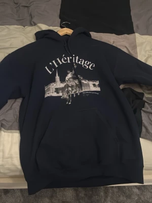 Mörkblå hoodie L'Héritage - (Buda på) Snygg mörkblå hoodie med tryck av L'Héritage och motiv från Stockholm. Stor känguruficka framtill och justerbar huva med dragsko. Perfekt för dig som gillar streetwear och unika prints.