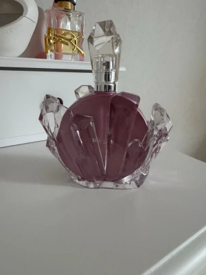 Ariana Grande parfym lila kristallflaska - Säljer en Ariana Grande parfym i en unik lila rund flaska med kristalliknande detaljer och genomskinligt lock. Flaskan är tillverkad av glas och har en cool, glittrig design som verkligen sticker ut på hyllan.