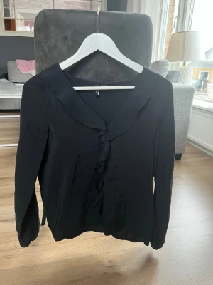 Svart blus med volangkant från Vero Moda - Stilren svart blus från Vero Moda med lång ärm och volangdetalj längs knäppningen framtill. 