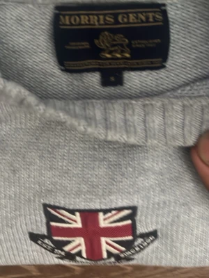 Grå tröja från Morris Gents - Snygg ljusgrå tröja från Morris Gents med Union Jack-detalj framtill. Tröjan har rund halsringning, ribbade muddar och nederkant. Perfekt för dig som gillar klassisk stil med brittisk touch.