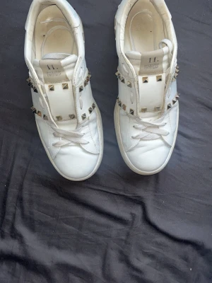 Vita Valentino sneakers med nitar - Snygga vita sneakers från Valentino med coola silverfärgade nitar runtom. Skorna är låga och har klassisk snörning. Tillverkade i läder med en chunky sula och unika detaljer som ger en edgy vibe. Perfekta för dig som vill sticka ut med din stil.