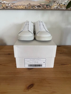 Vita sneakers från Oliver Cabell - Stilrena vita sneakers från Oliver Cabell i klassisk low-top modell. Skorna är tillverkade i slätt läder med vita snören och diskret branding. Perfekta för en clean och modern look. Snygg minimalistisk design som passar till det mesta.