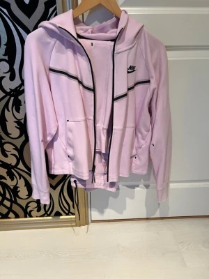 Ljusrosa hoodie och byxor från Nike - Ljusrosa hoodie från Nike med svart dragkedja och svarta detaljer längs bröstet. Ljusrosa byxor från Nike sälj i set. Hoodien har en huva och två fickor framtill. Perfekt för dig som gillar sportig och avslappnad stil. Materialet är mjukt och bekvämt, idealiskt för chill eller träning.