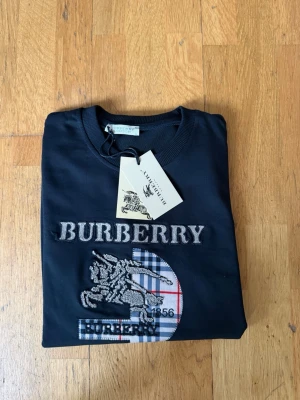 Svart sweatshirt från Burberry - Svart sweatshirt från Burberry med broderad logga och rutig detalj framtill. Tröjan har rund halsringning, långa ärmar och ribbade muddar. Klassisk och stilren design med Burberry-tryck även på ärmen.