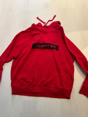 Röd hoodie från Gina Tricot - Röd hoodie från Gina Tricot med svart tryck där det står 'journey' i rött. Klassisk modell med huva och dragsko. Mjuk insida och ribbade muddar vid ärmslut och nederkant. Perfekt för en chill och avslappnad stil.