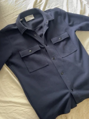 Mörkblå overshirt  - Stilren mörkblå overshirt från Weekday med två stora bröstfickor med lock och tryckknappar. Skjortkrage och knappstängning framtill. Perfekt lager på lager-plagg i mjukt material.