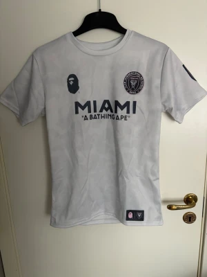 Vit MIAMI BAPE t-shirt - Snygg vit t-shirt från A Bathing Ape x Inter Miami med tryck fram och bak. MIAMI-logga och BAPE-apa framtill, samt stort BAPE 93-tryck på ryggen. Klassisk rund hals och korta ärmar. Perfekt för dig som gillar streetwear och fotbollsinspirerad stil.
