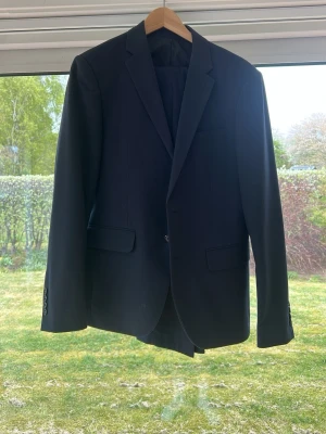 Svart klassisk kavaj med byxor till - Stilren svart kavaj med klassisk passform och enkla knappar framtill. Kavajen har två fickor med lock och en bröstficka. Perfekt för dig som vill ha en tidlös look till kostymen. Passar dig som är 175cm lång och väger ca 70-75kg 