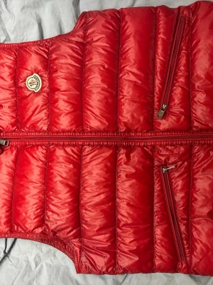 Moncler gui väst - Snygg röd dunväst från Moncler med quiltad design och två snedställda dragkedjefickor framtill. Västen har Monclers klassiska logga på bröstet och är tillverkad i glansigt material som ger en sportig vibe. Perfekt för lager-på-lager och streetstyle.