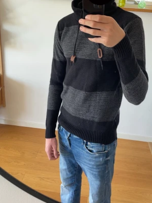 Svart och grå randig stickad hoodie - Helt ny, Stickad hoodie med breda svarta och grå ränder, hög krage med dragsko och ribbstickade muddar. Perfekt för dig som gillar en avslappnad men ändå snygg stil. Materialet är mjukt och värmande, passar bra till jeans. Inköpt i Spanien 2026 säljer pga den är för liten. 