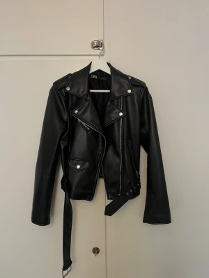 Svart skinnjacka från Zara - Snygg svart skinnjacka från Zara med klassisk bikerstil, silvriga dragkedjor och knappar. Jackan har bälte i midjan, fickor med lock och dragkedja samt axelklaffar. Perfekt för en cool och trendig look.