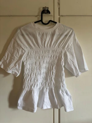 Vit blus med volang - Superfin vit blus med smockad midja och volang nertill. Blusen har korta ärmar och rund halsringning, vilket ger en söt och trendig look. Perfekt att styla med jeans eller kjol för en fräsch outfit.