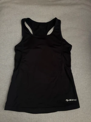 Svart träningstopp från SOC - Snygg svart träningstopp från SOC med brottarrygg och logga framtill. Tillverkad i ett lätt och stretchigt material som andas, perfekt för gymmet eller löpning. Sitter tajt och ger bra rörelsefrihet under träning.💞