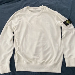Vit sweatshirt från Stone Island - Vit sweatshirt från Stone Island med klassisk rund hals och ribbade muddar. Tröjan har den ikoniska Stone Island-loggan på vänster ärm. Perfekt för en clean och stilren look.