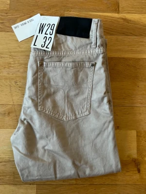 Beige chinos från Tiger of Sweden - Snygga beige chinos från Tiger of Sweden med klassisk rak passform och fem fickor. Byxorna har bälteshällor, knappgylf och är tillverkade i ett mjukt bomullsmaterial. Perfekta för en stilren och avslappnad look.