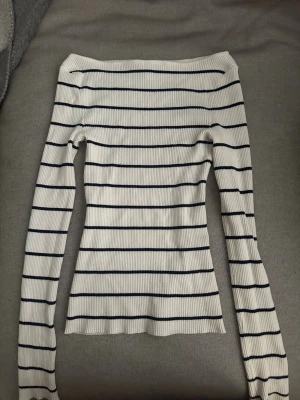 Randig ribbad topp från Amisu - Säljer en vit ribbad topp från Amisu med mörkblå horisontella ränder. Toppen har offshoulder-modell och långa ärmar, vilket ger en trendig och smickrande passform. Perfekt att styla med jeans eller kjol för en snygg look.