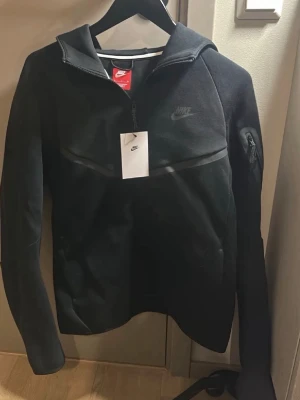 Svart Nike hoodie med dragkedja - Snygg svart hoodie från Nike med halv dragkedja framtill och diskret Nike-logga på bröstet. Hoodien har huva, lång ärm och en liten ficka med dragkedja på ena ärmen. Tillverkad i mjukt och bekvämt material, perfekt för en sportig och avslappnad stil.