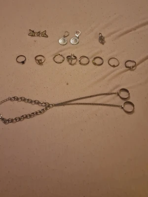 Set med smycken - Säljer ett set med tre örhängen i form av fjärilar. Örhängena är små och har en vit färg med guldfärgade kanter. De är tillverkade i metall och har en söt och lekfull design som passar perfekt för dig som gillar unika accessoarer. Det finns även flera olika ringar, ett armband med ringar på. Ja, du får allt i bilden för detta pris.