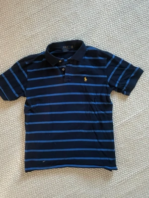 Randig pikétröja från Polo Ralph Lauren - Mörkblå pikétröja med blå ränder från Polo Ralph Lauren. Klassisk krage, korta ärmar och broderad gul logga på bröstet. Perfekt för en avslappnad men ändå stilren look.
