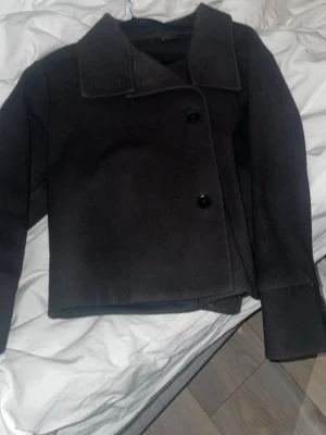 Svart kort rock med bred krage - Stilren svart rock med bred krage och två stora knappar framtill. Modellen är kortare och har raka ärmar. Perfekt för dig som gillar en klassisk och enkel look. Materialet ser ut att vara ullblandning. Skriv för mer bilder