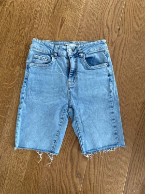 Ljusblå jeansshorts med rå kant - Snygga ljusblå jeansshorts med hög midja och rå, fransig kant nertill. Klassisk femficksmodell med dragkedja och knapp framtill. Från Ginatricot i storlek 36.