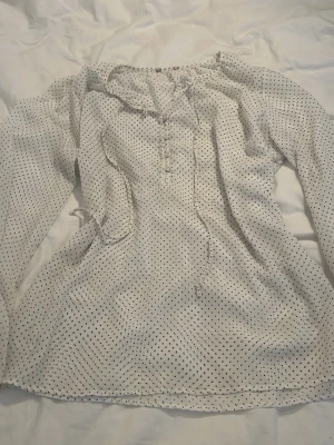 Vit prickig blus med volang - Söt vit blus med svarta små prickar och volangdetaljer vid halsringningen. Blusen har knytband framtill och långa ärmar. Tunt och luftigt material som ger en avslappnad och trendig look.