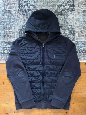 Marinblå Ralph Lauren Cardigan/Hybrid Zip Hoodie - Hej! 👋 Säljer en riktigt snygg quiltad Ralph Lauren hybrid zip jacka / cardigan i storlek L. Sjukt lyxigt och stilrent plagg! Tröjan är i gott skick, men har blivit lite naturligt blekt på armbågarna – inget som stör helhetsintrycket. 100% äkta med originaltvättlappar kvar och en fullt fungerande QR-kod för smidig verifiering ✅ Perfekt lyxplagg för kyligare kvällar eller som övergångsjacka! Fraktar snabbt! 💬🚛📦 