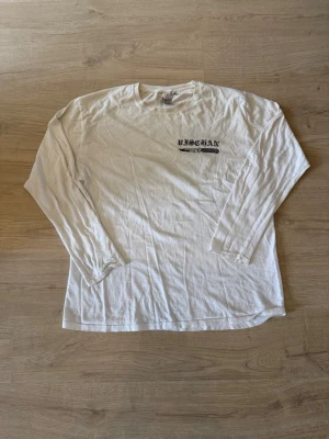 Vit långärmad t-shirt med tryck - Vit långärmad t-shirt med svart tryck på bröstet och ett stort grafiskt motiv på ryggen. Skön passform och tillverkad i mjuk bomull, perfekt för en avslappnad stil.