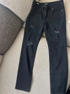 Svarta skinny jeans Jack & Jones - Svarta skinny jeans från Jack & Jones, modell Liam. Jeansen har slitna detaljer på benen och klassisk femficksdesign. Snygg passform med smala ben och normal midja. Perfekta för en trendig och avslappnad stil.
