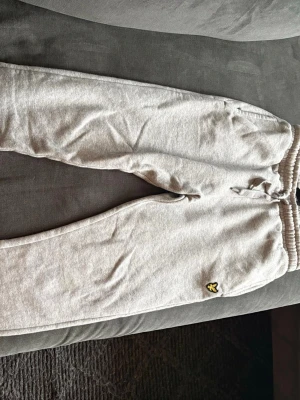 Grå mjukisbyxor från Lyle & Scott - Snygga grå mjukisbyxor från Lyle & Scott med resår och dragsko i midjan. Byxorna har en klassisk passform och är tillverkade i mjukt bomullsmaterial. Liten gul logga på benet ger en stilren touch.