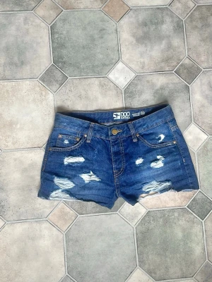 Blå lågmidjade jeansshorts med slitningar från DOO - Snygga blå jeansshorts från DOO Australia med slitna detaljer och klassisk femficksdesign. Shortsen har normal passform, knapp och dragkedja framtill samt råa benslut för en trendig look. Perfekta för varma dagar och enkel att matcha med olika toppar.